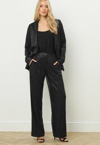 Zwarte paillettenpak met een op maat gemaakte blazer en wijde broekspijpen. Gladde stof met glanzende pailletten, gecombineerd met een zwarte top en hoge hakken.