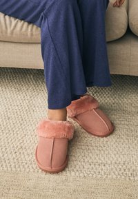 Chaussons en daim rose avec un bord en fourrure rose pelucheux, ornés d'un motif cousu. Portés avec un pantalon bleu marine sur un tapis beige.