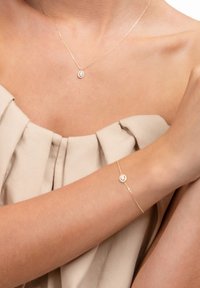 Gold necklace with circular pendant and matching bracelet, featuring smooth chain and small circular accents, displayed against beige fabric.