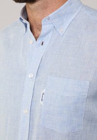 Camicetta a quadri blu chiaro con colletto, taschino e tessuto testurizzato. Presenta bottoni bianchi e una piccola etichetta del marchio sulla tasca.