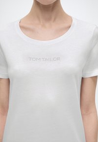Γυναίκα που φοράει λευκό T-shirt με στρογγυλή λαιμόκοψη και το κείμενο "TOM TAILOR" φτιαγμένο από μικρές στρας στο στήθος.