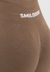 Braune Leggings mit gerippter Textur und einem weißen "SMILODOX"-Logo am Bund, im Nahaufnahme gezeigt.