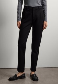 Zwarte op maat gemaakte broek met een gladde textuur, met een recht pootontwerp en een subtiele vouw. Gepaard met zwarte instappers en een grijze top.