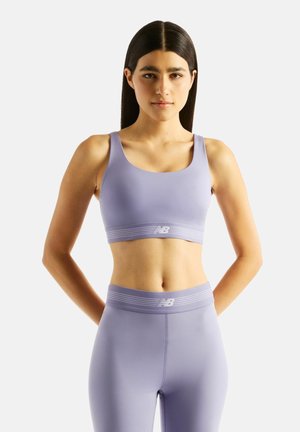 Frau steht mit den Armen hinter dem Rücken, trägt einen passenden helllila Sport-BH und Leggings mit New Balance-Logo und blickt nach vorn.