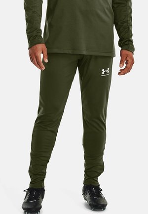 Mand iført olivengrønne Under Armour sportbukser og matchende top, stående med sorte sportssko på hvid baggrund.