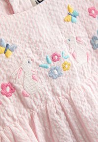 Svetloružové bavlnené šaty so zvislými pruhmi; zdobené vyšívanými zajacmi, kvetmi a motýľmi v pastelových farbách. Detail volánov na leme.