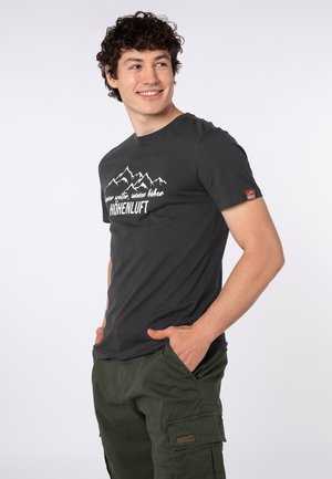 Young man with curly hair wearing a black mountain-themed T-shirt and green cargo pants, smiling and looking to the side.