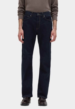 Mand iført mørkeblå straight-leg jeans, brune loafers og en taupefarvet langærmet sweater, stående foran en hvid baggrund.