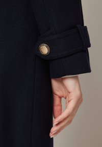 Tali navy płaszcza wełnianego z mankietem i złotym guzikiem. Materiał wydaje się teksturowany, co podkreśla ogólny design.