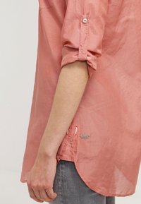 Ljusrosa långärmad skjorta med uppkavlade ärmar, med detaljer som knappar och justerbar dragsko vid fållen, tillverkad av ett smidigt tyg.