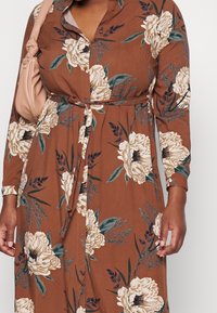 Kvinde iført en brun floral kjole med store hvide blomster, med knyttet taljebælte, som bærer en lys pink håndtaske over skulderen.