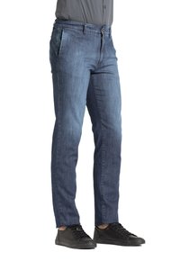 Jeans in denim blu scuro con vestibilità slim, dotati di chiusura con zip, tasche anteriori e posteriori, e leggere sfumature sul tessuto. Abbinati a sneakers nere.