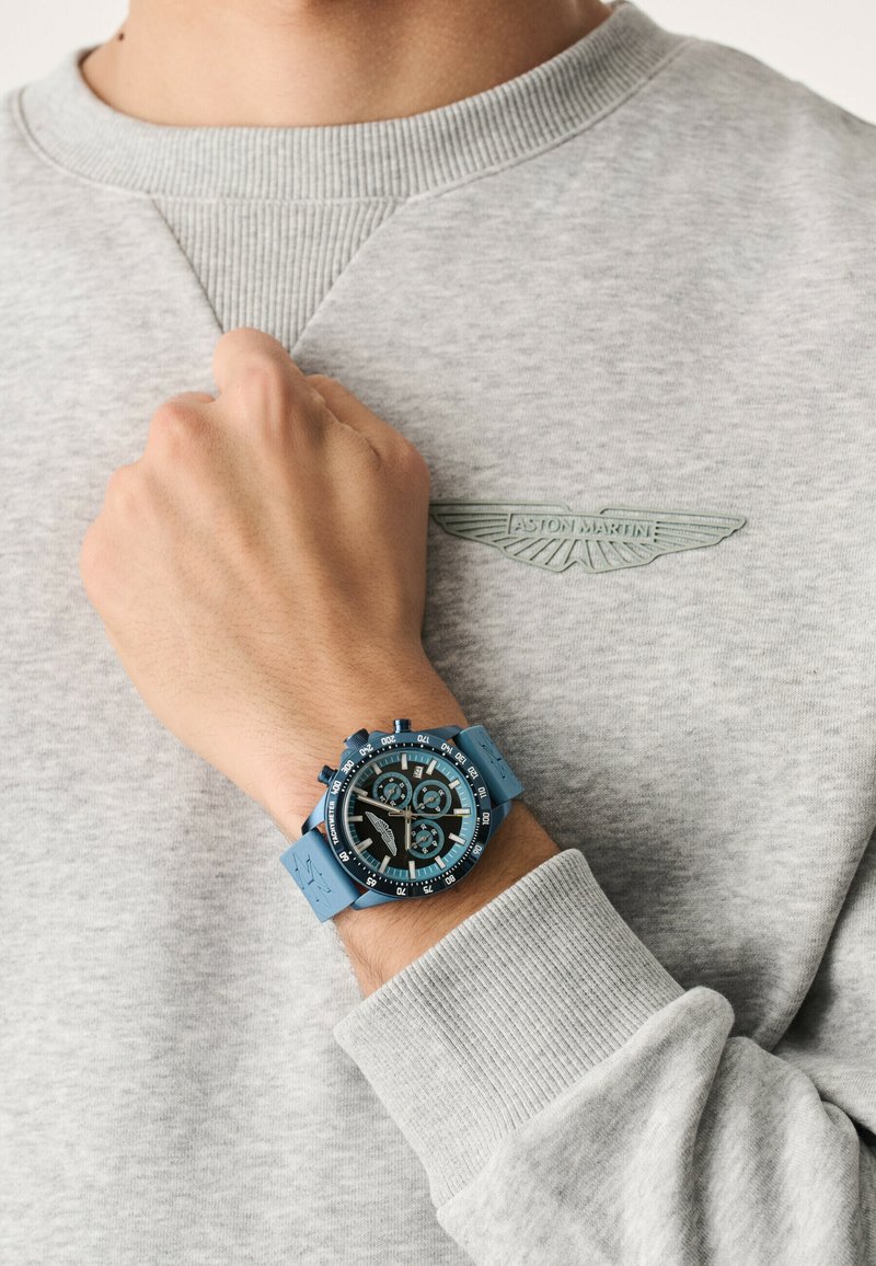 Blauer Chronograph mit rundem Zifferblatt, Gummiband und schwarzen Akzenten, getragen an einer Hand vor einem hellgrauen Sweatshirt mit Logo.