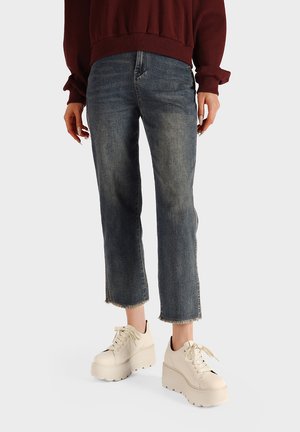 Persona che indossa jeans blu sbiaditi corti, un maglione color bordeaux e sneakers bianche chunky con zeppa, in piedi su uno sfondo semplice.