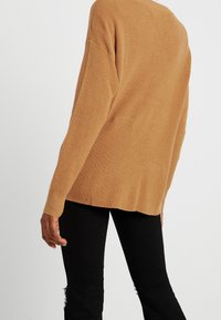 Długi, prążkowany sweter w jasnobrązowym kolorze, z luźnym krojem i lekko zaokrąglonym dołem, zestawiony z czarnymi, postrzępionymi dżinsami.