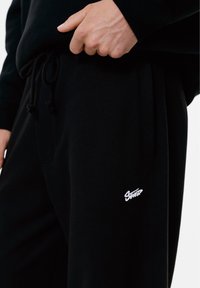 Zwarte katoenen joggers hebben een verstelbare drawstring tailleband en een klein wit logo. De stof heeft een gladde, zachte textuur.