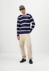 Polo à manches longues rayé bleu marine et blanc avec col, associé à un pantalon beige et des chaussures noires, porté par un mannequin.