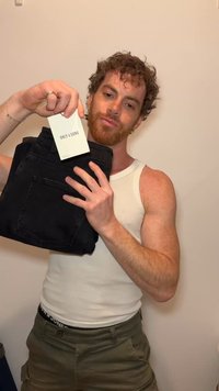 Άνδρας με σγουρά μαλλιά και γένια κρατάει μαύρα τζιν με ετικέτα «ONLY & SONS», φορά λευκό αμάνικο πουκάμισο και ελιάς παντελόνι.