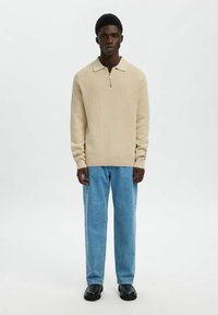 Beżowy sweter na suwak o strukturze dzianiny, w połączeniu z jasnoniebieskimi jeansami o prostych nogawkach i czarnymi botkami. Gładkie tło.