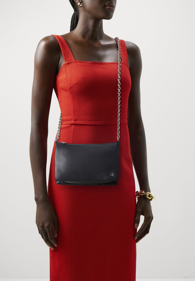 Borsa a tracolla in pelle nera con una tracolla a catena, caratterizzata da una finitura liscia e un discreto logo. Indossata sopra un vestito rosso con texture.