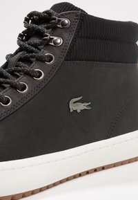 Svarta högskor med slät yta, med silverfärgade öglor, texturerad manschett och en vit gummimikro. Lacoste-logotyp på sidan.