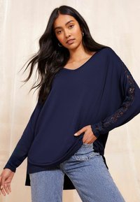 Navy oversized V-neck majica od lagane tkanine ima duge rukave s čipkastim detaljima, u kombinaciji s svijetlim trapericama.