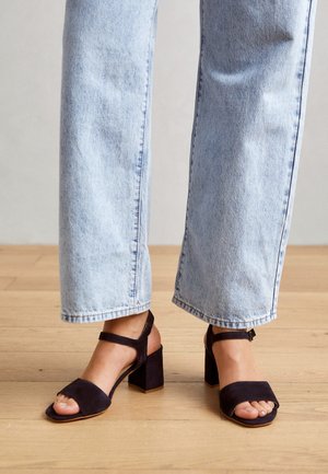 Fødder iført sorte ruskindssandaler med blokhæl og ankelremme, kombineret med lyseblå jeans med lige ben på et trægulv.