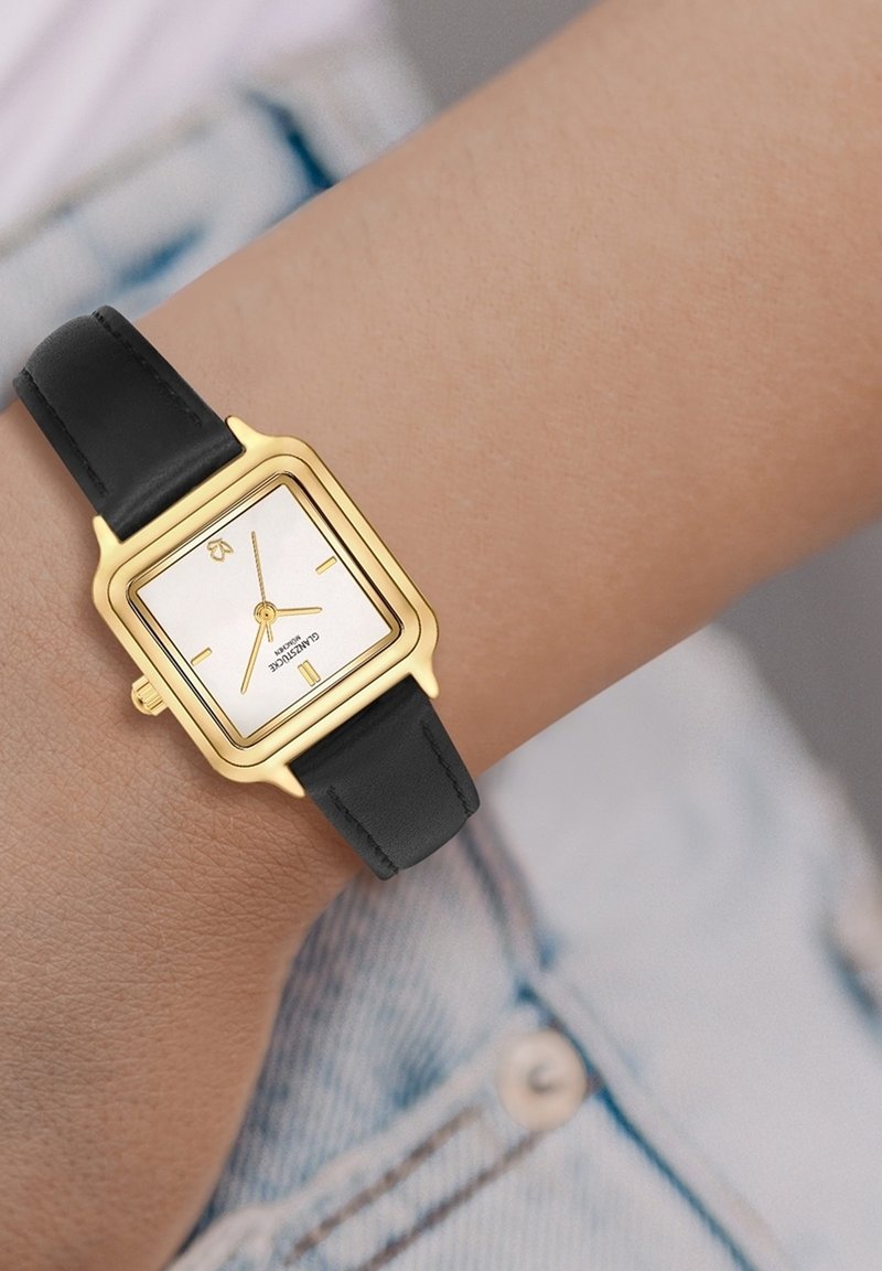 Gold square watch with a white dial, black leather strap, gold accents, and minimalist hour markers. Visible crown on the side.