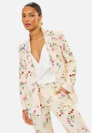 Woman wearing a cream floral suit with pink, red, and blue flowers, paired with a white deep V-neck top and gold hoop earrings.