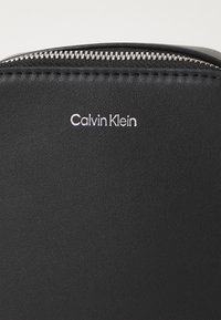 Svart syntetisk lommebok med strukturert overflate, med sølvglinsende glidelås og preget "Calvin Klein"-logo på framsiden.