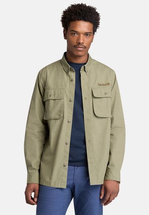 Mann trägt ein olivgrünes Hemd über einem marineblauen T-Shirt und blauen Hosen, steht vor einem einfarbigen hellen Hintergrund.