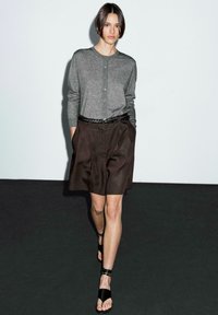 Femme marchant vêtue d'un cardigan gris à boutons, de shorts plissés marron avec une ceinture tressée et de sandales noires à lanières autour de la cheville sur fond uni.