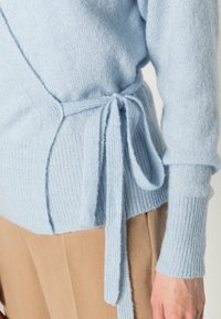 Jasnoniebieski sweter dziany z zapinanym designem, z wiązaniem w talii i ściągaczami na mankietach. Miękka tekstura, dopasowane rękawy.