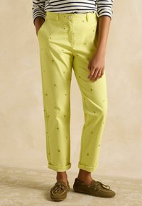 Kerged kollased püksid, millel on tikitud lillemustrid, sirge lõikega ja üles keeratud sääred, koos pruunide sussidega, neutraalse tausta ees.