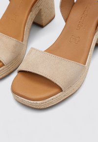 Sandalias de tacón bloque de ante beige con una amplia tira en la parte delantera. Presentan una textura suave y lisa, y un acento de cuerda trenzada alrededor de la suela.