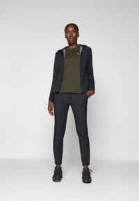 Black zip-up jacket with textured sleeves, olive green t-shirt, dark pants, and black athletic shoes. Simple design, fitted silhouette.