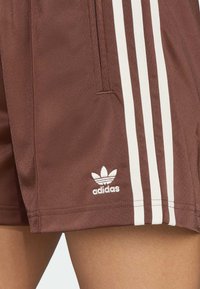 Calções atléticos castanhos com três riscas brancas ao longo do lado e um logótipo da Adidas bordado na bainha. Tecido suave e leve.