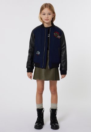 Veste d'aviateur marine avec des manches en cuir noir, ornée de patchs brodés. Assortie à une jupe verte et des bottes lacées noires.