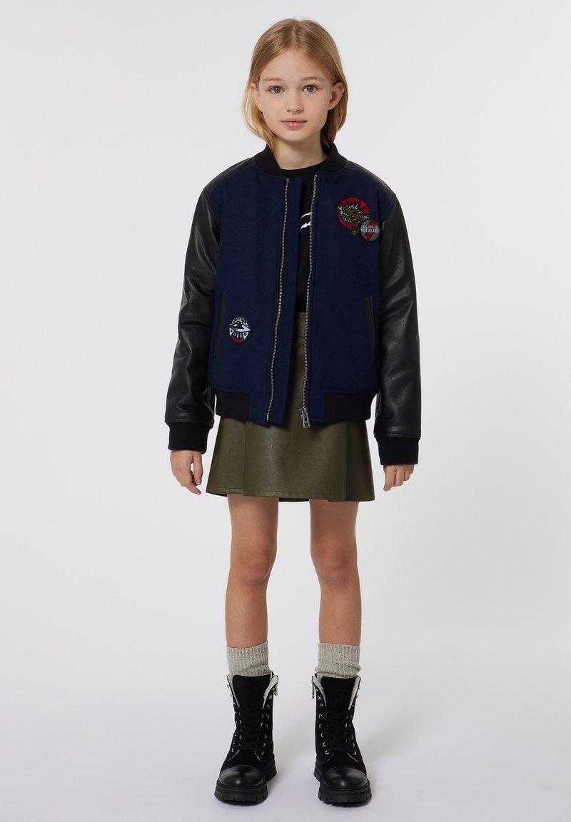 Veste d'aviateur marine avec des manches en cuir noir, ornée de patchs brodés. Assortie à une jupe verte et des bottes lacées noires.