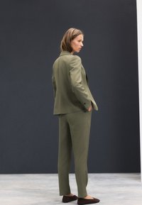 Oļīvu zaļš kostīms ar strukturētu žaketi un taisnu kāju biksēm, kombinēts ar brūnām slip-on kurpēm. Gluda tekstūra un minimāls dizains.