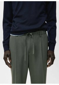 Hombre con suéter azul oscuro y pantalones verde oliva con cinturón de tela anudado, de pie con las manos relajadas a los lados.