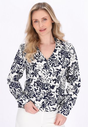 Szőke nő laza hullámos hajjal, fekete-fehér virágos, hosszú ujjú gombos ingben és fehér nadrágban, egyszínű háttér előtt állva.