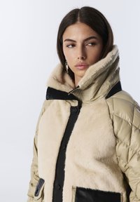 Jeune femme aux cheveux bruns portant une veste matelassée beige avec un grand col en fourrure et des accents en cuir noir, regardant sur le côté.
