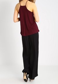 Burgundowa bezrękawnik o luźnym fasonie, zestawiony z czarnymi spodniami o szerokich nogawkach i butami na wysokim obcasie, podkreślający prosty design i kontrast kolorystyczny.
