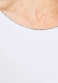 Tessuto bianco con una texture a coste, caratterizzato da una dettagliata catena argentata lungo il collo arrotondato. Il tono della pelle è visibile sotto il tessuto.