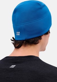 Persoon met kort krullend haar die een felblauwe gebreide muts draagt en een zwart shirt met een klein reflecterend logo op de achterkant.