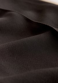 Prim-plan al unui material maro închis, cu țesătură nervurată, textură țesută vizibilă și pliuri delicate.