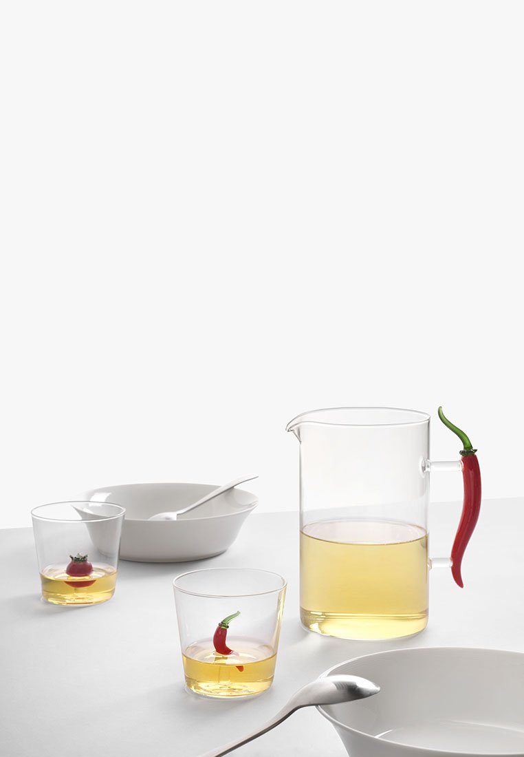 Przezroczysty dzbanek z uchwytem w kształcie czerwonej papryki, dwie szklanki z czerwoną papryką w cieczy, białe miski i łyżka na płaskiej powierzchni.