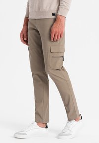 Pantaloni cargo beige con vestibilità slim, dotati di tasche laterali e una tasca con zip. Indossati con sneakers bianche per un look informale.