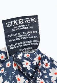 Marineblaue Stoff mit einem weißen und roten Blumenmuster, versehen mit einem Pflegeetikett mit Waschhinweisen und Symbolen in der Nähe des Ausschnitts.